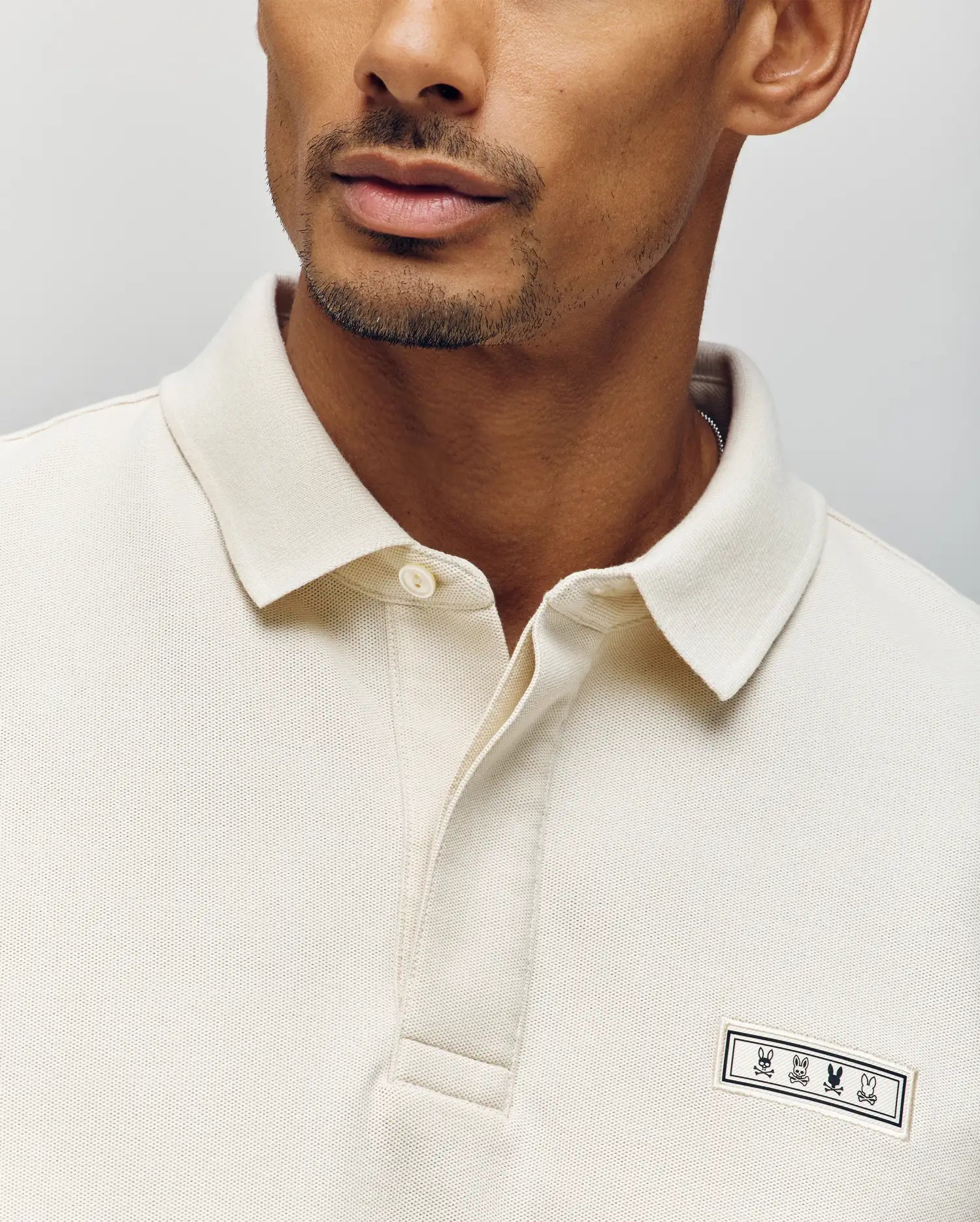 A man wears the Psycho Bunny MENS AXTON PIQUE POLO (MT0100011) in cream Pima cotton, featuring a concealed button placket and a jacquard logo patch with three minimalistic animal icons. The image shows only his lower face and upper torso.