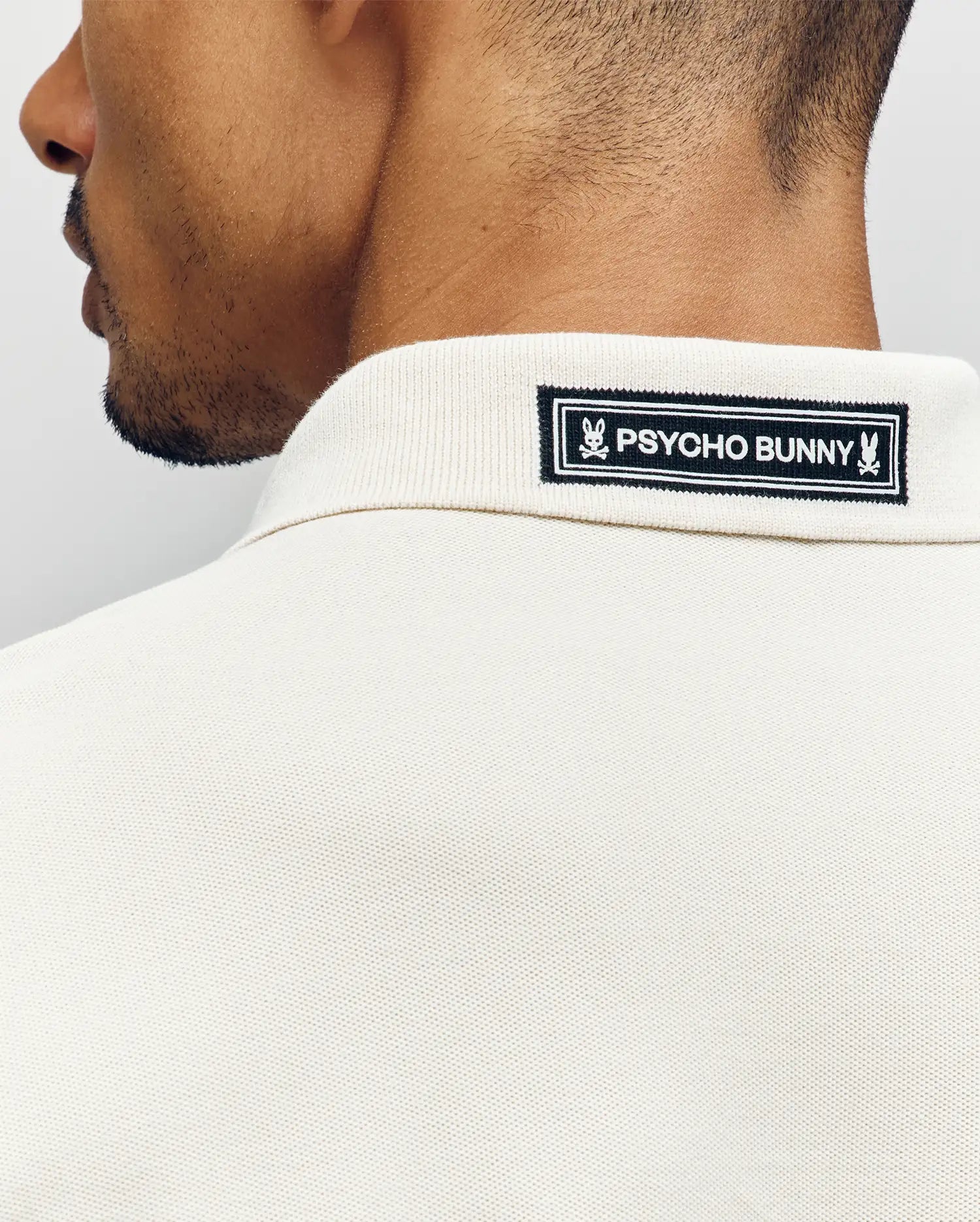 Close-up of the back of a man's neck and shoulders wearing the Psycho Bunny MENS AXTON PIQUE POLO (MT0100011) in cream, featuring a black-and-white jacquard-woven logo patch sewn onto the collar.