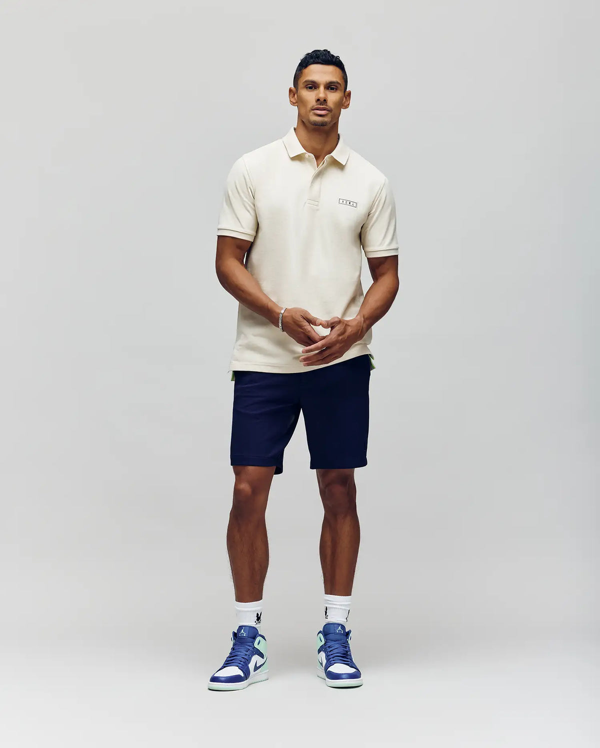 A man stands against a plain background wearing the Psycho Bunny MENS AXTON PIQUE POLO - MT0100011 in cream, paired with navy shorts, white socks, and white and blue sneakers. He faces forward with his hands clasped in front of him.