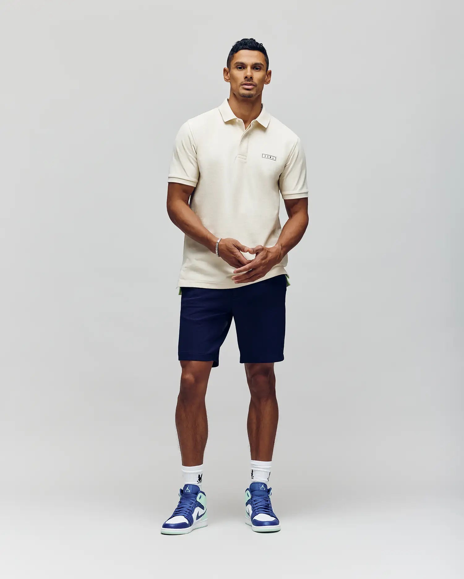 A man stands against a plain background wearing the Psycho Bunny MENS AXTON PIQUE POLO - MT0100011 in cream, paired with navy shorts, white socks, and white and blue sneakers. He faces forward with his hands clasped in front of him.
