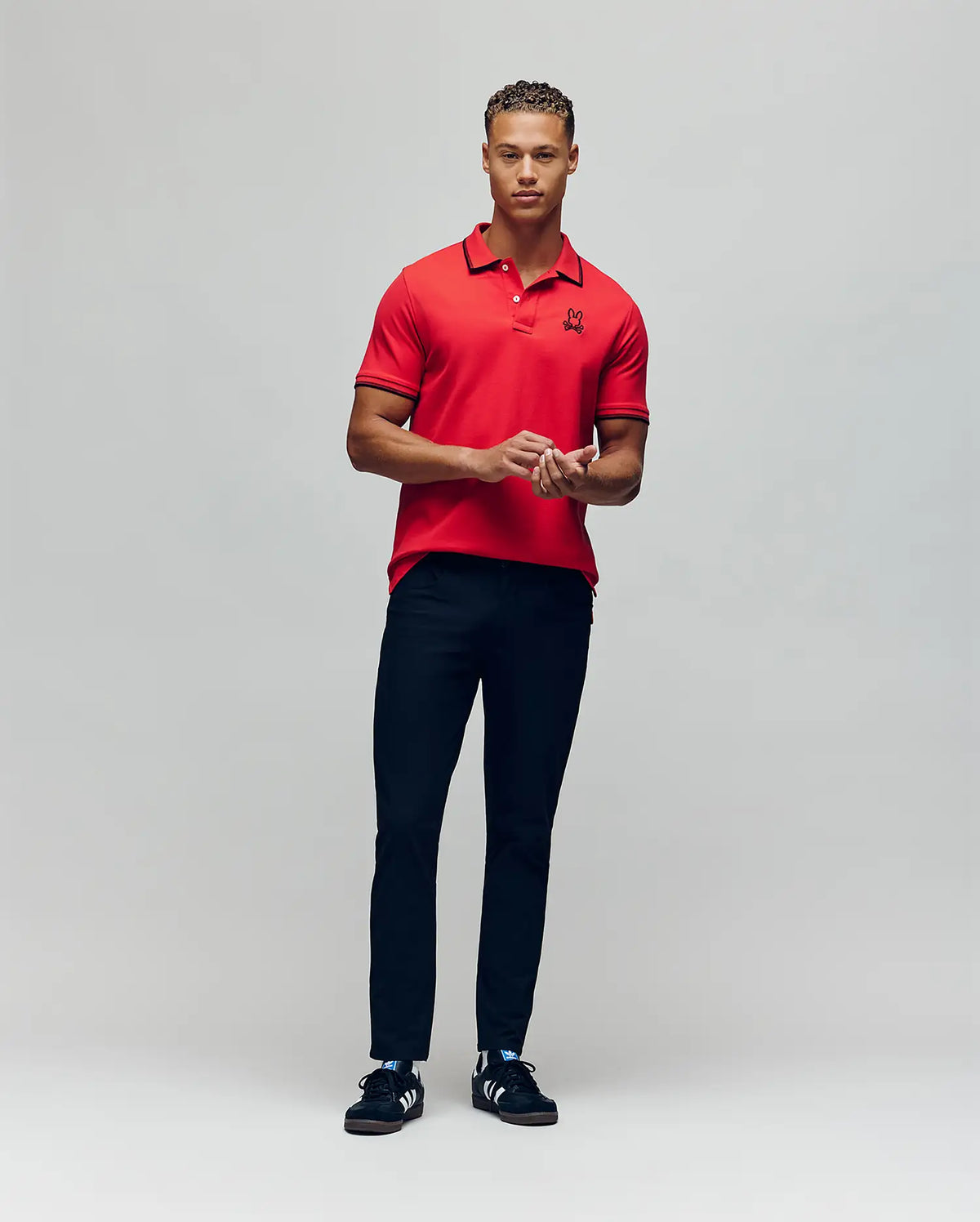 A man stands against a plain background wearing the Psycho Bunny MENS JUDE PIQUE POLO - MT0100012 in red. He faces forward with his hands together and looks aside, showing off the signature embroidered Bunny logo.