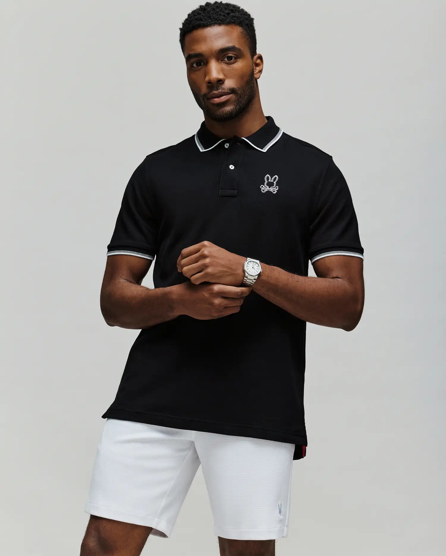 A man stands with arms crossed in white shorts against a plain background, wearing the Psycho Bunny MENS JUDE PIQUE POLO - MT0100012, crafted from soft Pima Cotton and featuring the brand's embroidered bunny logo on the chest.