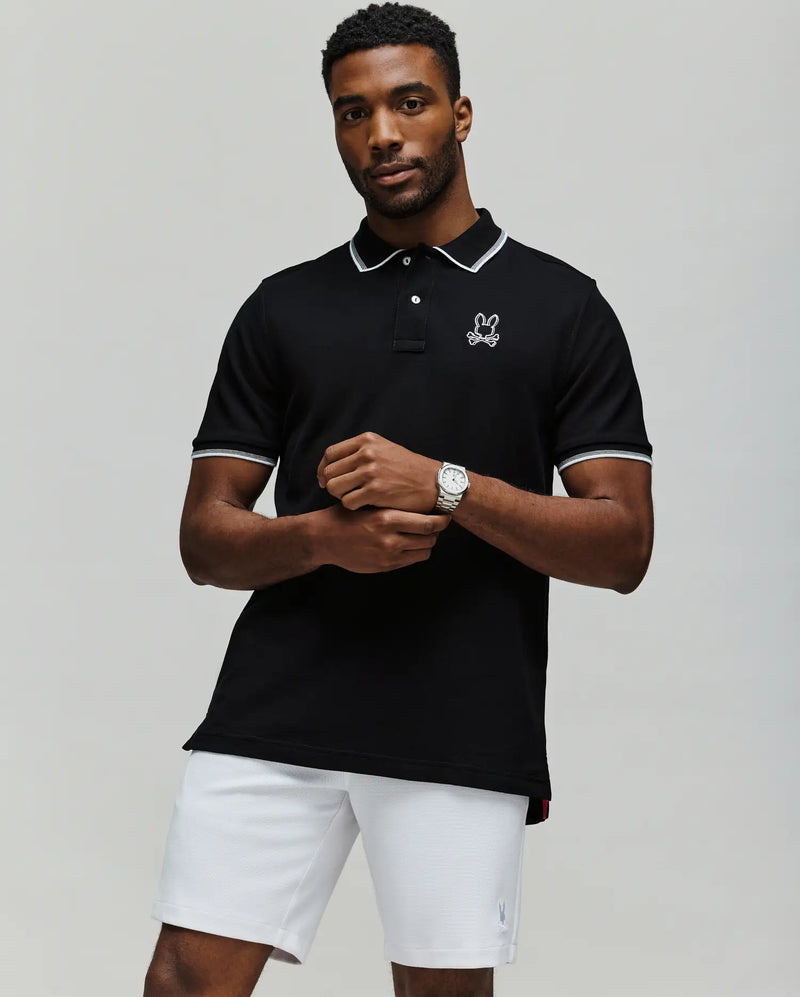 A man stands with arms crossed in white shorts against a plain background, wearing the Psycho Bunny MENS JUDE PIQUE POLO - MT0100012, crafted from soft Pima Cotton and featuring the brand's embroidered bunny logo on the chest.