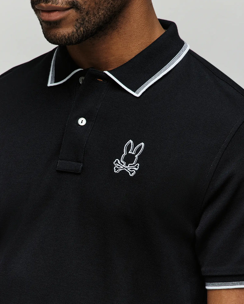A man wears the Psycho Bunny MENS JUDE PIQUE POLO (MT0100012), a black Pima cotton polo with white collar trim and an embroidered bunny with crossbones logo on the left chest.