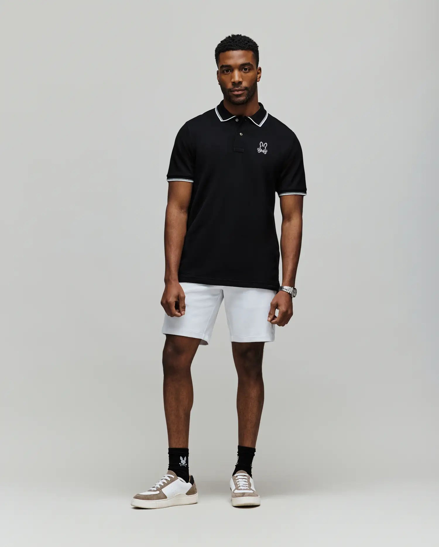 A man stands facing forward in a Psycho Bunny MENS JUDE PIQUE POLO - MT0100012, paired with white shorts, black socks, and white sneakers, against a plain light gray background.