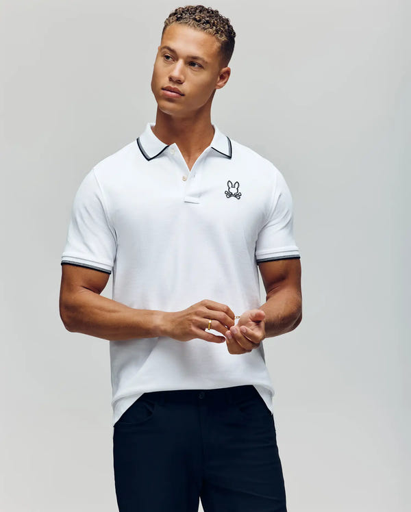 A man wears the Psycho Bunny MENS JUDE PIQUE POLO (MT0100012), a white Pima cotton shirt with black trim and an embroidered Bunny logo on the chest, standing against a plain background looking slightly to the side.
