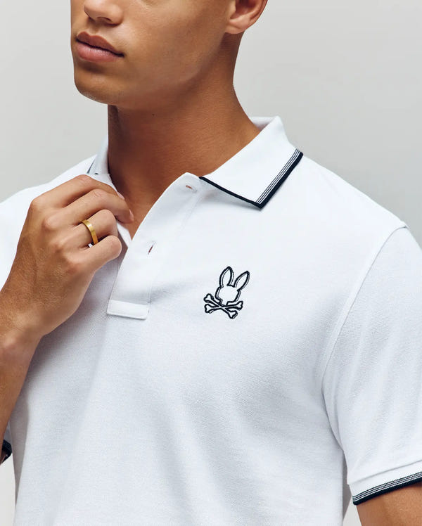 A person wearing the Psycho Bunny MENS JUDE PIQUE POLO (MT0100012), made from soft Pima cotton with black trim and an embroidered Bunny logo above crossed bones, touches the collar while sporting a gold ring.