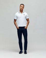 A man stands against a light background, wearing the Psycho Bunny MENS JUDE PIQUE POLO - MT0100012 in white, featuring an embroidered Bunny emblem, paired with black pants and shoes. His hands are in his pockets as he faces the camera.