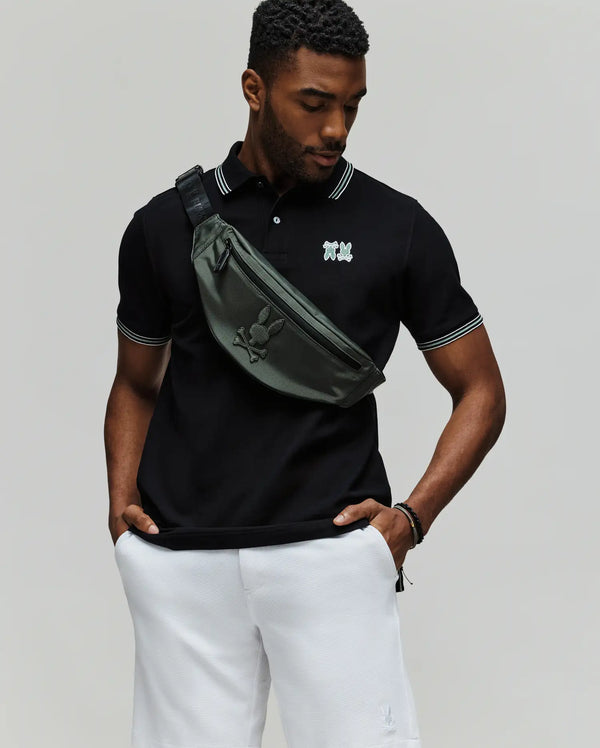 A man stands against a plain background wearing the Psycho Bunny MENS ARCHIVE NEON BUNNY POLO (MT0100018) in black with white trim, paired with white shorts and a green crossbody bag. The shirt features an embroidered Bunny detail. He looks down, hands in pockets.