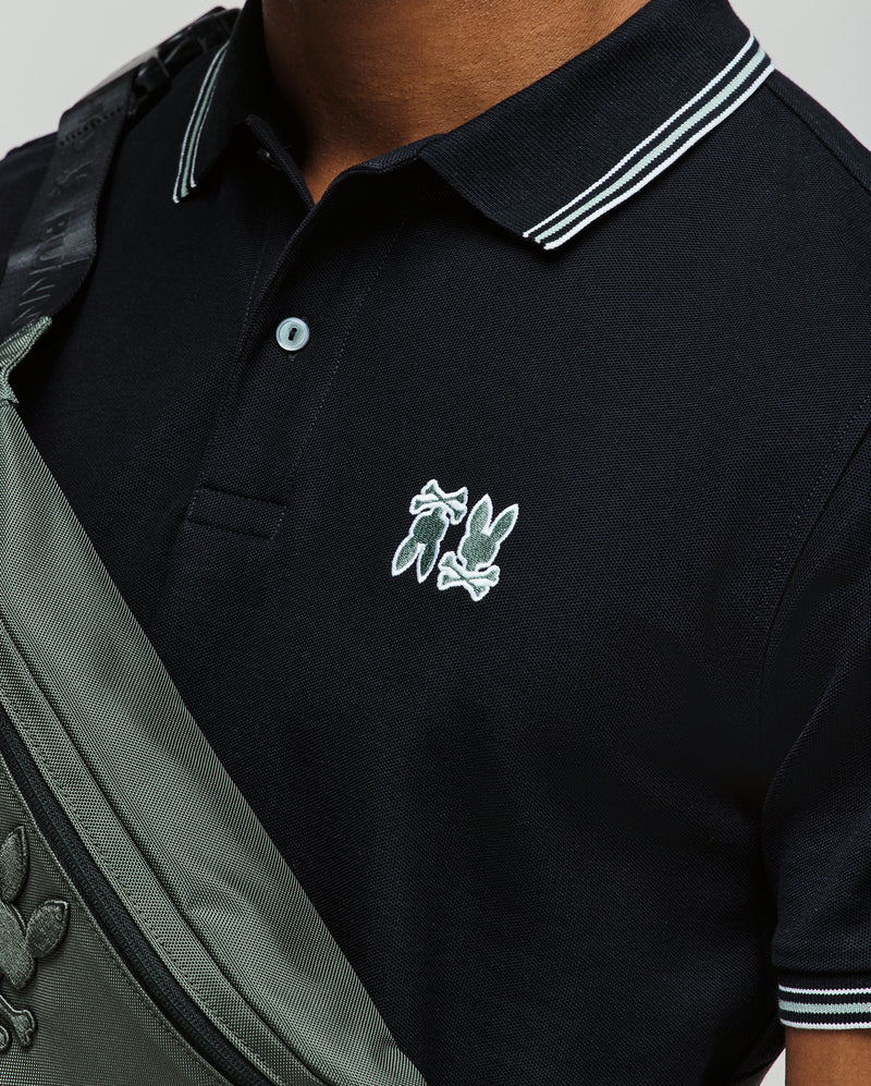 A person wearing a Psycho Bunny MENS ARCHIVE NEON BUNNY POLO (MT0100018) in black with white trim and an embroidered Bunny logo, paired with a grey crossbody bag featuring the same logo. Only the upper torso is visible.