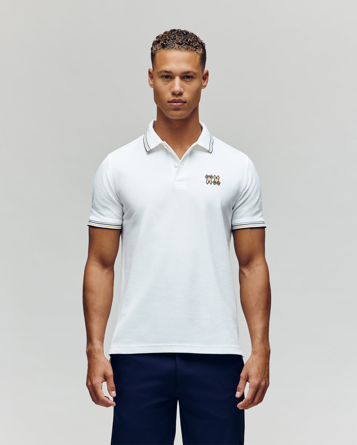 A man faces forward in dark pants and a Psycho Bunny MENS ARCHIVE NEON BUNNY POLO MT0100018—a regular fit white, 100% Pima cotton short-sleeve shirt featuring a small embroidered Bunny on the chest—against a plain light background.