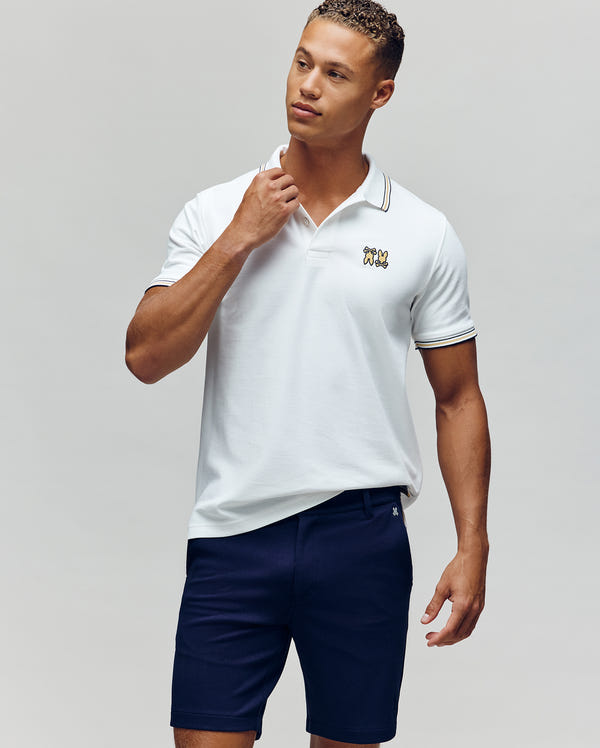 A man stands against a plain background in the Psycho Bunny MENS ARCHIVE NEON BUNNY POLO - MT0100018, a white, regular fit 100% Pima cotton shirt with an embroidered Bunny logo, paired with navy blue shorts. He looks to the side and touches his collar.