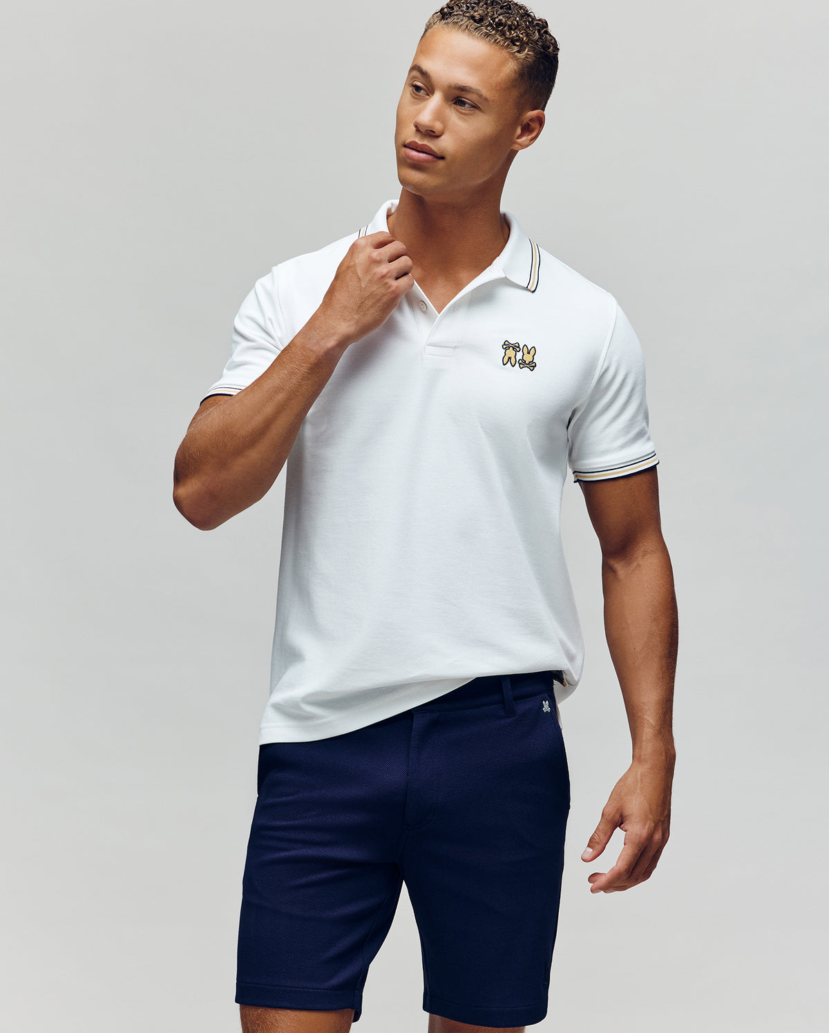 A man stands against a plain background in the Psycho Bunny MENS ARCHIVE NEON BUNNY POLO - MT0100018, a white, regular fit 100% Pima cotton shirt with an embroidered Bunny logo, paired with navy blue shorts. He looks to the side and touches his collar.