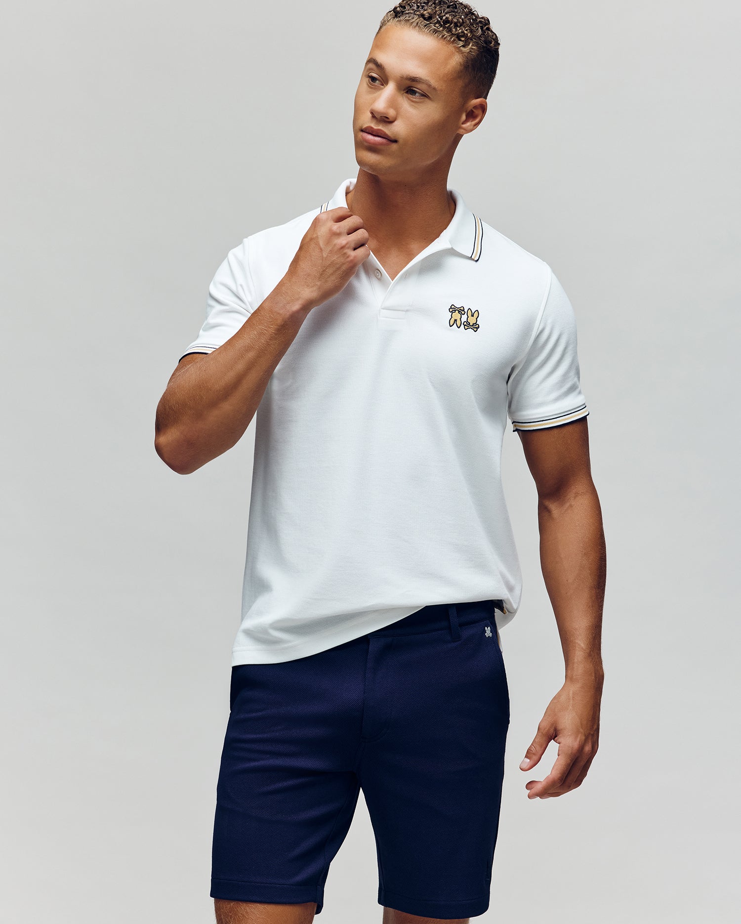 A man stands against a plain background in the Psycho Bunny MENS ARCHIVE NEON BUNNY POLO - MT0100018, a white, regular fit 100% Pima cotton shirt with an embroidered Bunny logo, paired with navy blue shorts. He looks to the side and touches his collar.