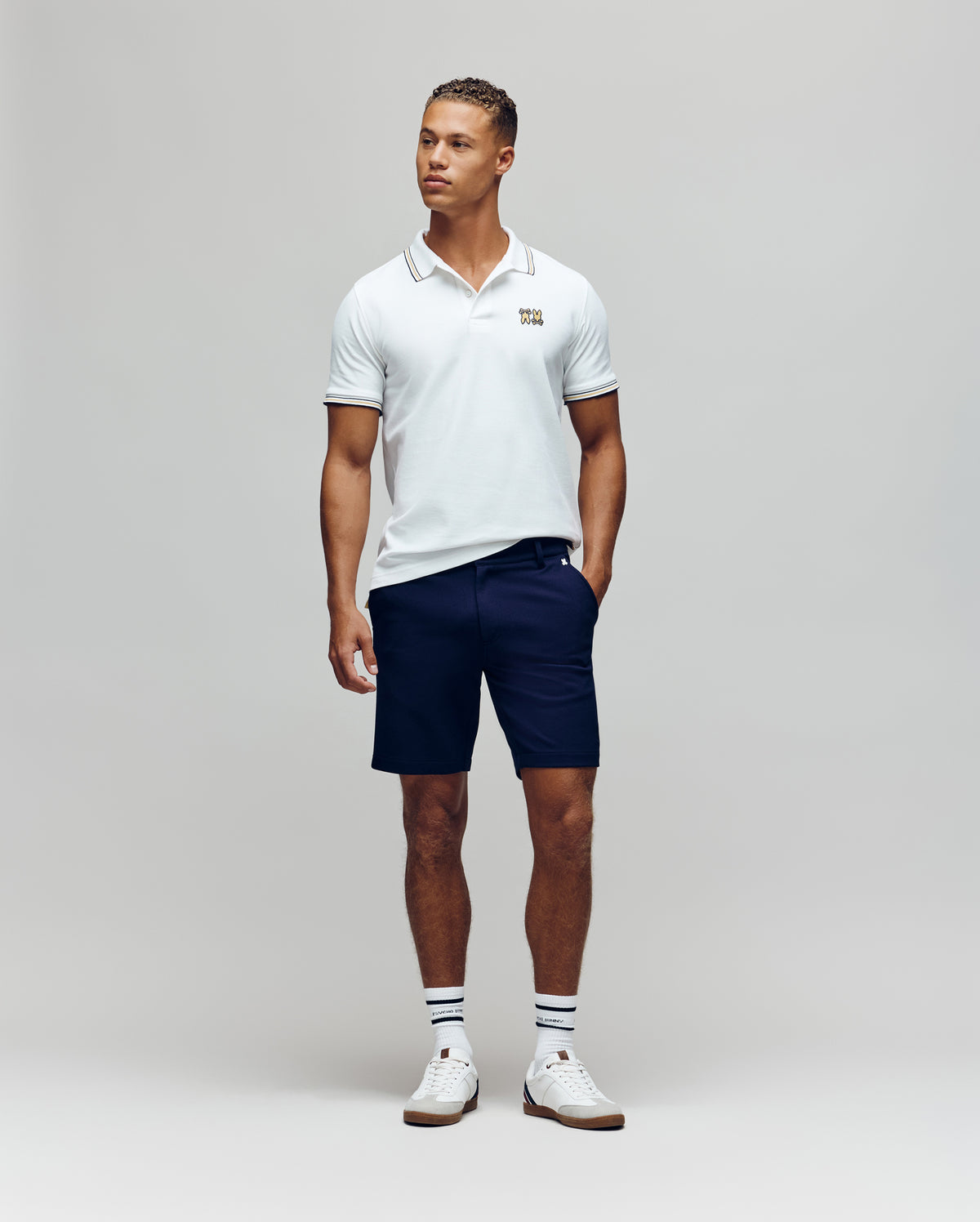 A man stands against a plain background wearing the Psycho Bunny MENS ARCHIVE NEON BUNNY POLO - MT0100018 in white, paired with navy shorts, striped white socks, and white sneakers. He has one hand in his pocket and looks slightly to the side.