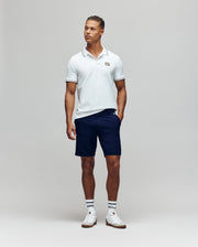 A man stands against a plain background wearing the Psycho Bunny MENS ARCHIVE NEON BUNNY POLO - MT0100018 in white, paired with navy shorts, striped white socks, and white sneakers. He has one hand in his pocket and looks slightly to the side.