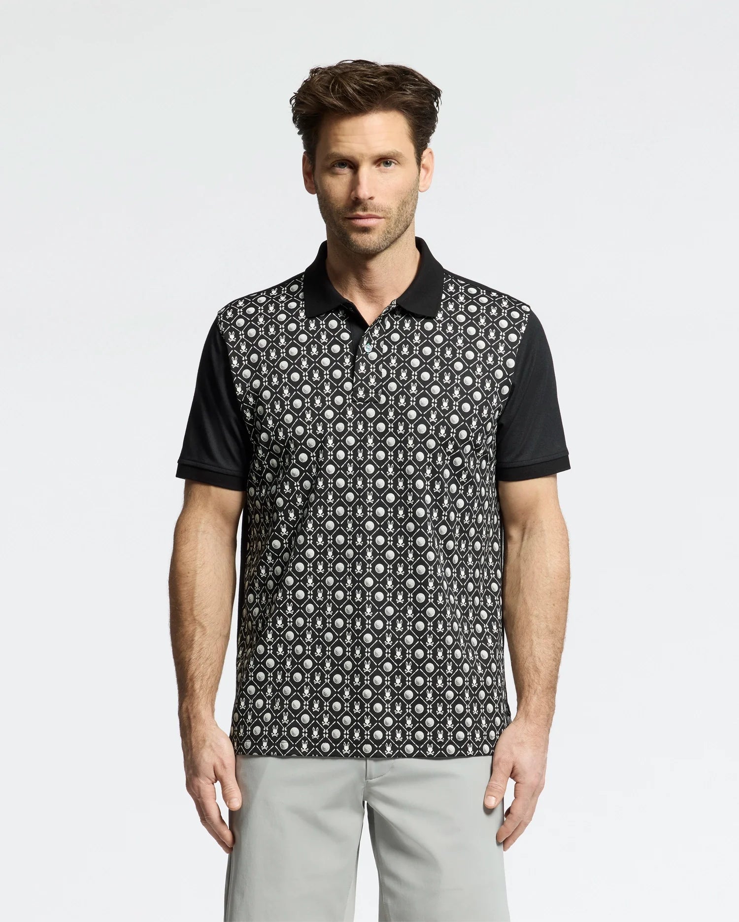 A man models the Psycho Bunny MENS BECKETT PIQUE POLO - MT0100022, featuring a black and white geometric front with solid black sleeves and collar. Against a white backdrop, he pairs it with light gray pants, exuding casual elegance.