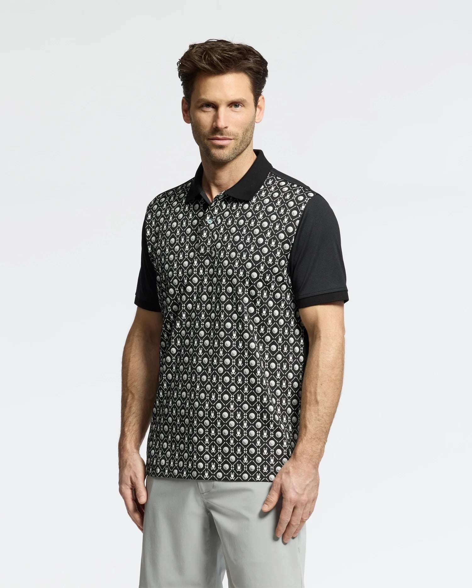 A man is wearing the Psycho Bunny MENS BECKETT PIQUE POLO - MT0100022, featuring a black and white golf-ball pattern with short sleeves, paired with gray pants, against a plain white background.