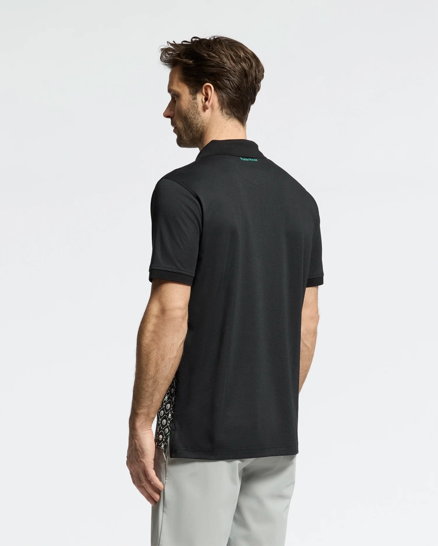 A man with brown hair is seen from behind, wearing gray pants and the Psycho Bunny MENS BECKETT PIQUE POLO - MT0100022, a black Pima cotton polo featuring a golf-ball-patterned design on the side panel, against a plain light background.