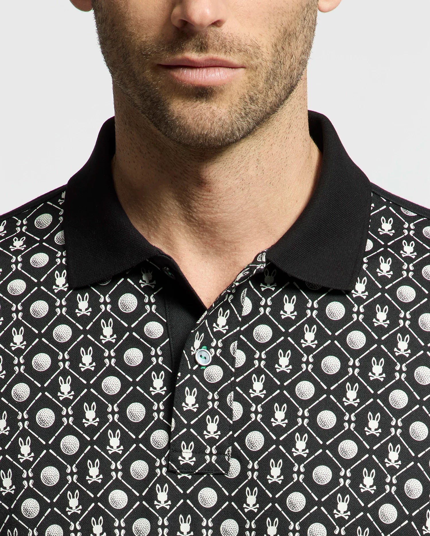 A man wears the Psycho Bunny MENS BECKETT PIQUE POLO, MT0100022, featuring a black Pima cotton base with playful white geometric patterns like golf-ball-style circles and abstract bunny motifs. It includes a black collar contrasting against its detailed design on a plain white background.