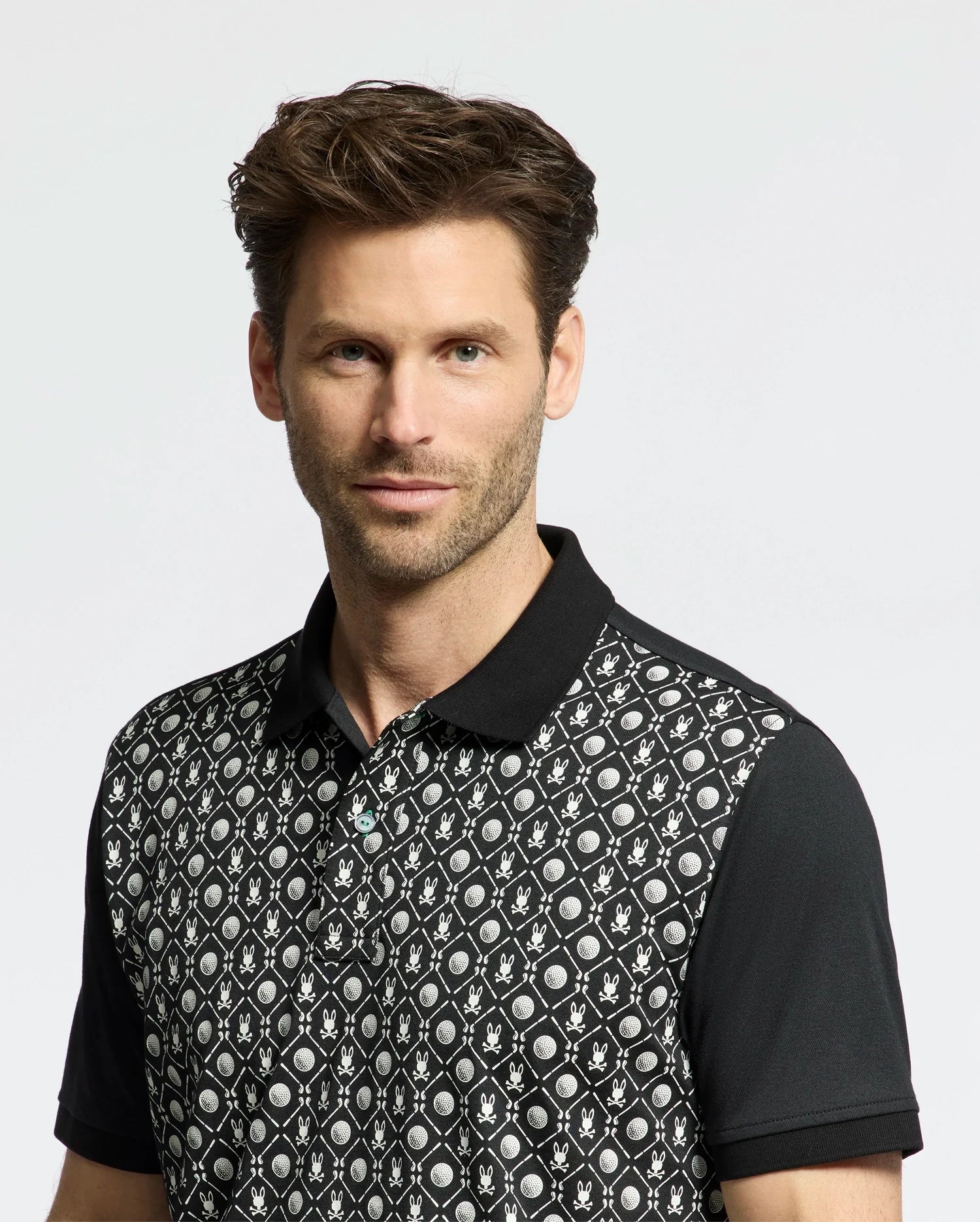 A man with short brown hair and a trimmed beard stands against a white background, wearing the Psycho Bunny MENS BECKETT PIQUE POLO - MT0100022. The Pima cotton polo with geometric patterns and floral designs, along with its four-way stretch, enhances his slight smile perfectly.
