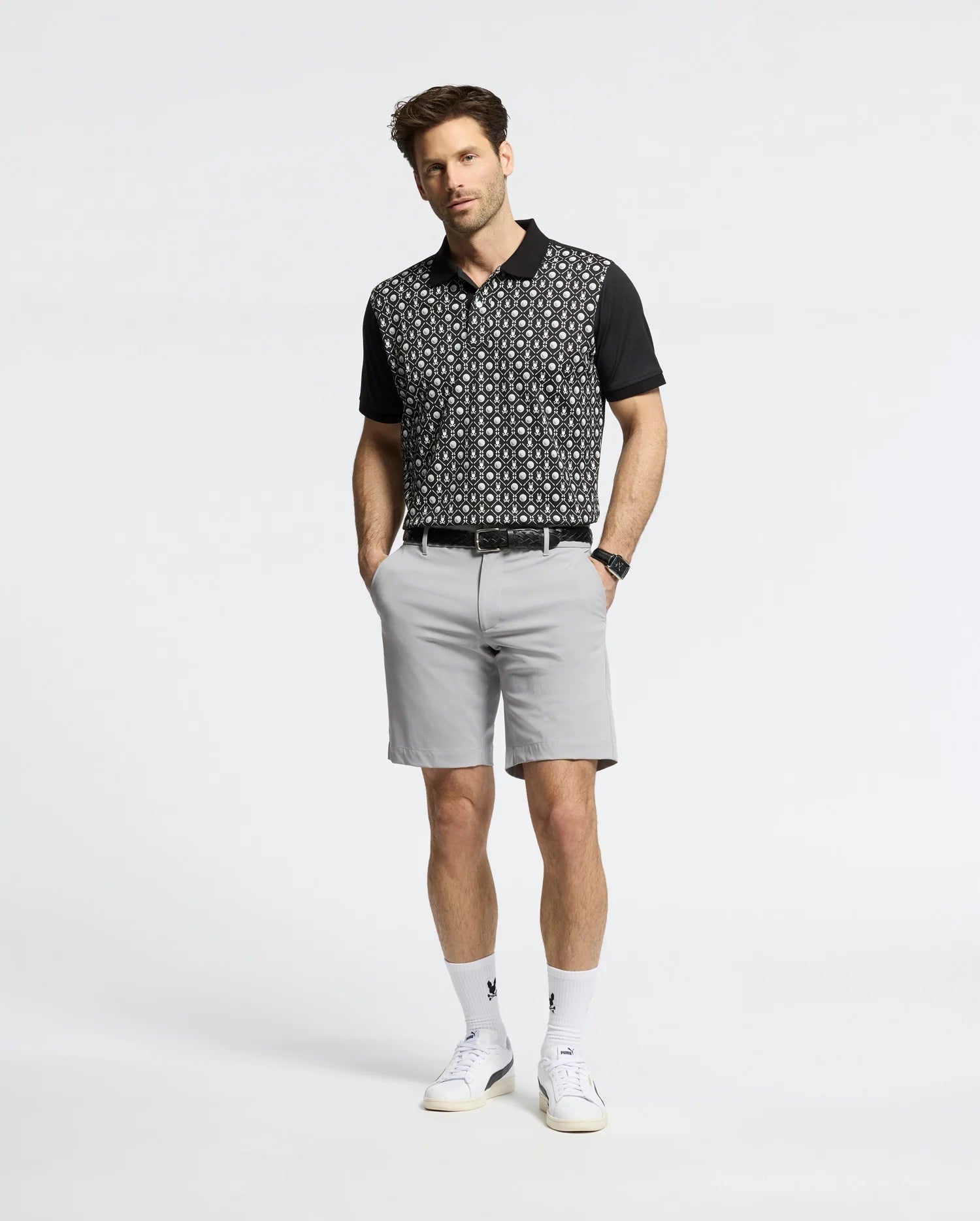 A man stands with hands in his pockets, wearing a Psycho Bunny MENS BECKETT PIQUE POLO (MT0100022), light gray shorts, white socks, and white sneakers against a plain light background.