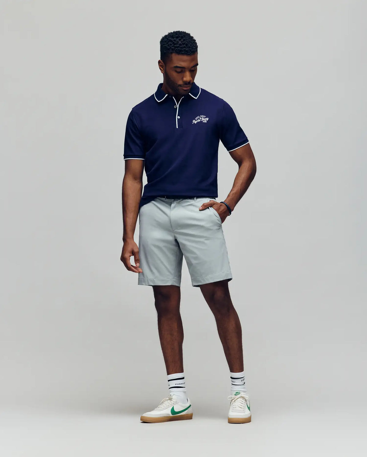 A man stands against a plain backdrop wearing the Psycho Bunny MENS MATTEO PIQUE POLO - MT0100024 in navy with white trim, paired with light gray shorts, white socks featuring green stripes, and white sneakers accented in green. He looks down, one hand in his pocket.