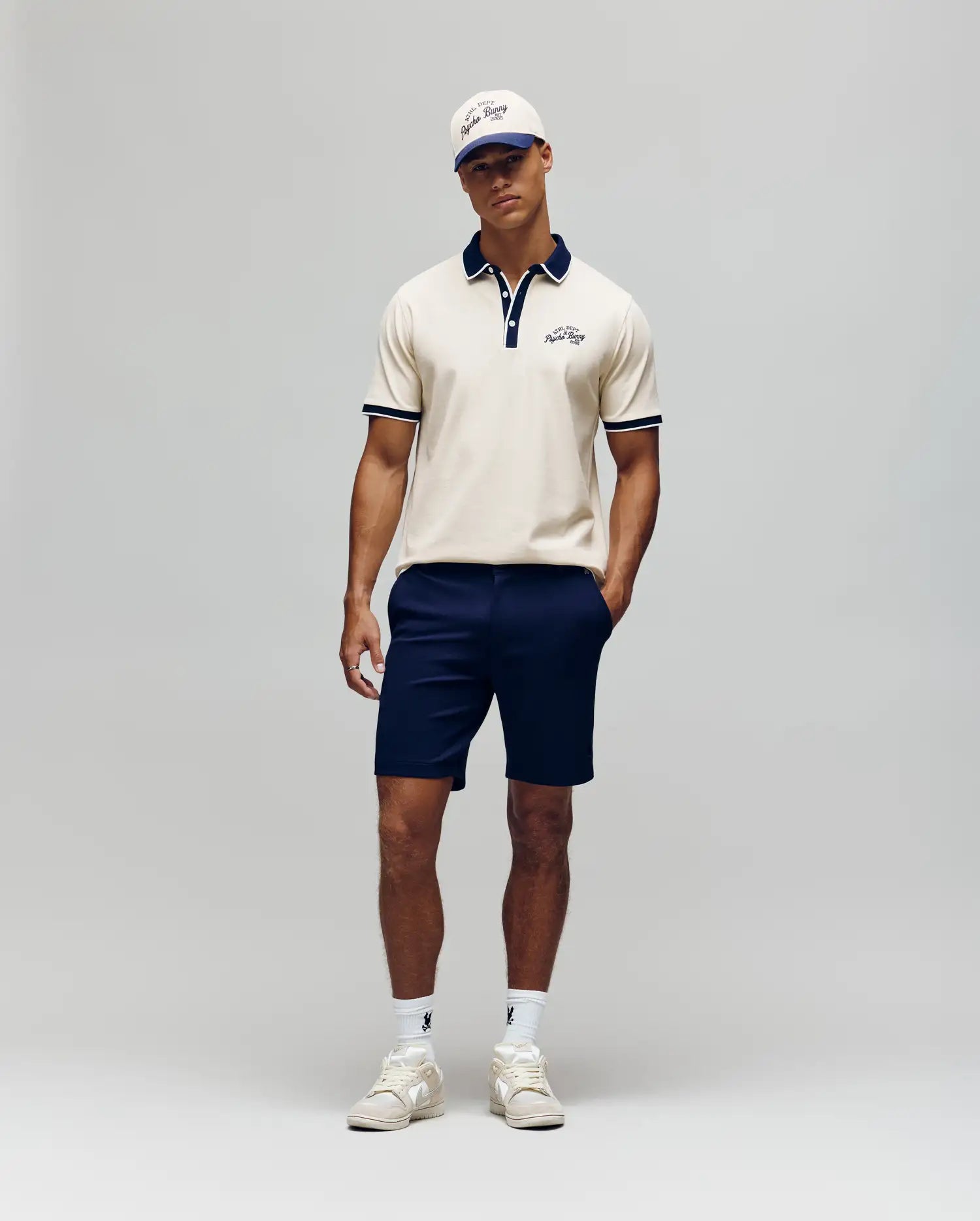 A man stands against a plain background wearing the Psycho Bunny MENS MATTEO PIQUE POLO - MT0100024, paired with navy shorts, white sneakers, a white cap, and sunglasses. His hands are in his pockets and he looks relaxed.