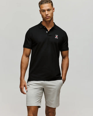 A man stands against a plain background wearing the Psycho Bunny MENS MISCHIEF BUNNY PIQUE POLO - MT0100025 in black with a small embroidered logo on the chest, paired with light gray shorts and his left hand in his pocket.