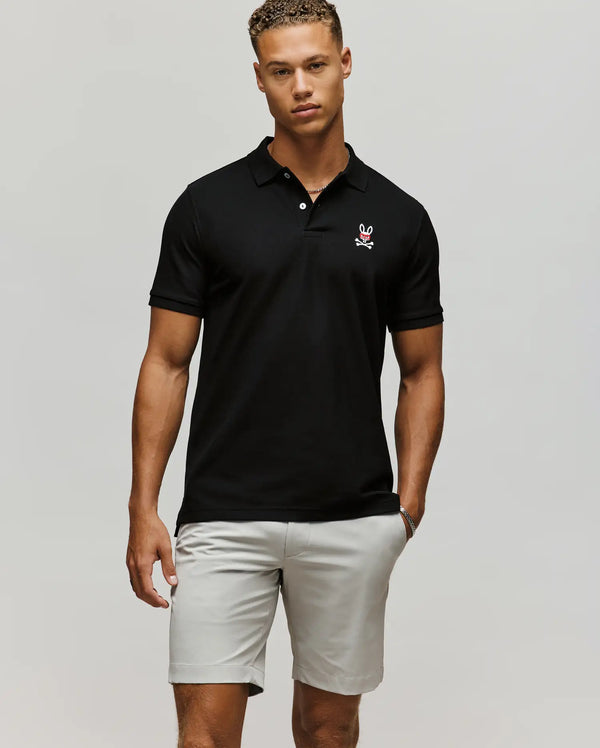 A man stands against a plain background wearing the Psycho Bunny MENS MISCHIEF BUNNY PIQUE POLO - MT0100025 in black with a small embroidered logo on the chest, paired with light gray shorts and his left hand in his pocket.