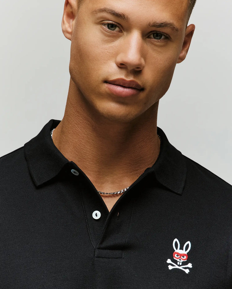 A man wears the Psycho Bunny MENS MISCHIEF BUNNY PIQUE POLO (MT0100025), a black collared shirt with an embroidered white bunny and crossbones logo on the chest, standing against a plain light background.