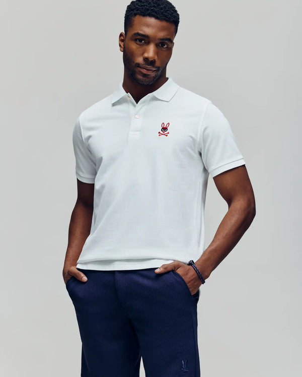 A man stands with hands in pockets, wearing a Psycho Bunny MENS MISCHIEF BUNNY PIQUE POLO - MT0100025 in white Pima cotton with a red embroidered chest logo, paired with dark blue pants against a light gray background.
