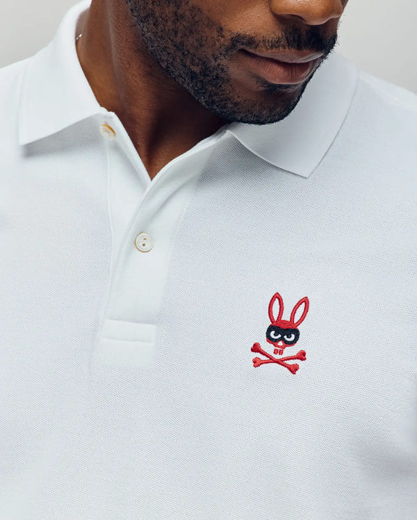 A man wears the Psycho Bunny Mens Mischief Bunny Pique Polo (MT0100025), made from soft Pima cotton, featuring a red and black embroidered bunny head and crossbones logo on the left chest.