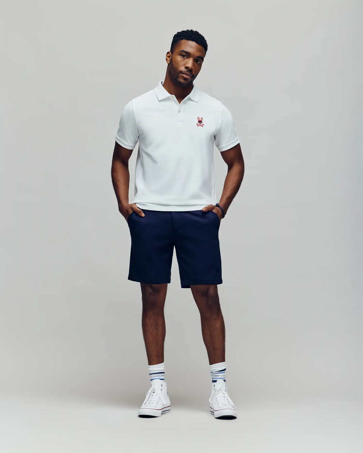 A man stands against a plain background wearing a white Psycho Bunny Mens Mischief Bunny Pique Polo (MT0100025) with a small red emblem, navy shorts, white socks with blue stripes, and white high-top sneakers. He looks relaxed with hands in pockets.