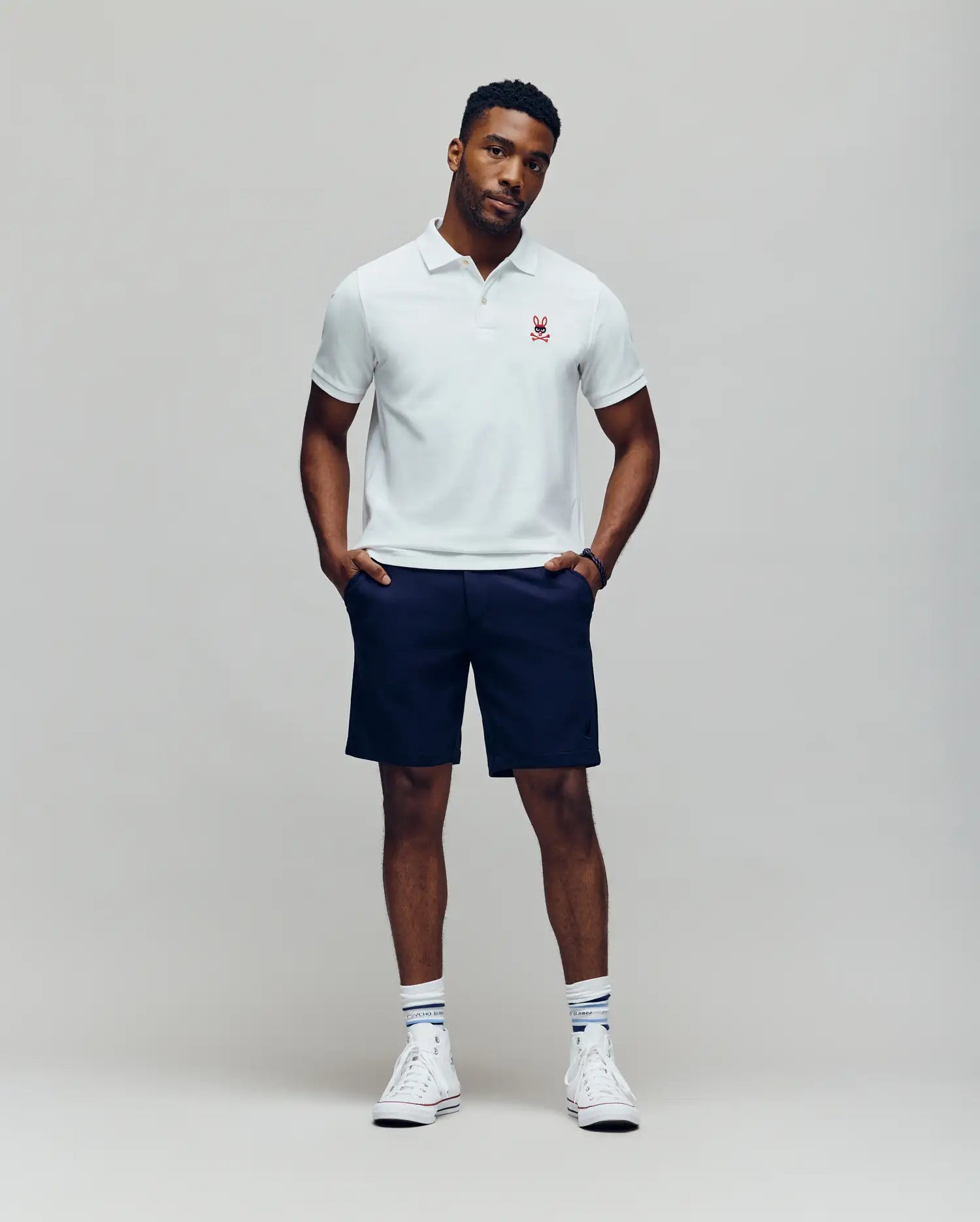 A man stands against a plain background wearing a white Psycho Bunny Mens Mischief Bunny Pique Polo (MT0100025) with a small red emblem, navy shorts, white socks with blue stripes, and white high-top sneakers. He looks relaxed with hands in pockets.