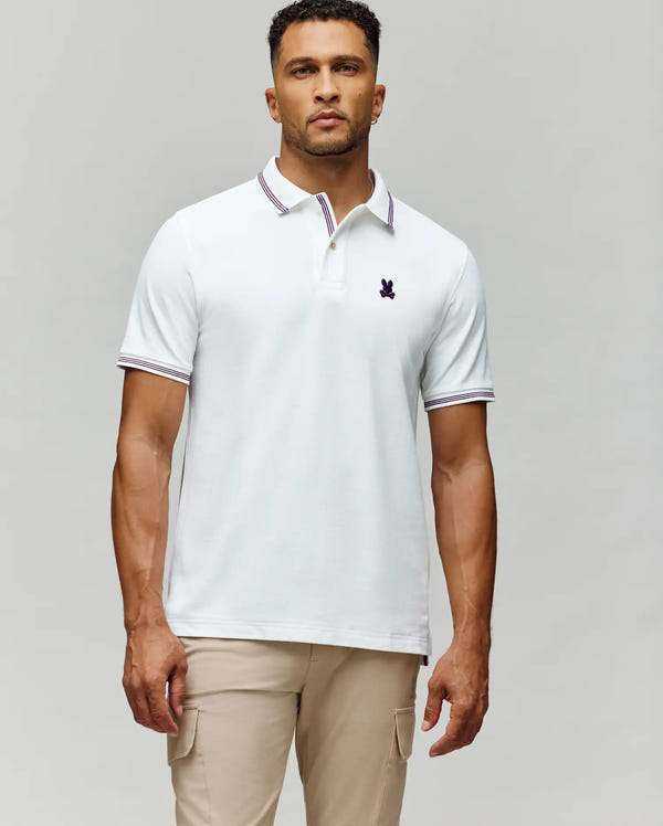 A man stands against a plain light gray background wearing beige pants and a Psycho Bunny MENS HART PIQUE POLO SHIRT (MT0100032), featuring white Pima cotton, an embroidered logo, plus striped collar and sleeve trim.