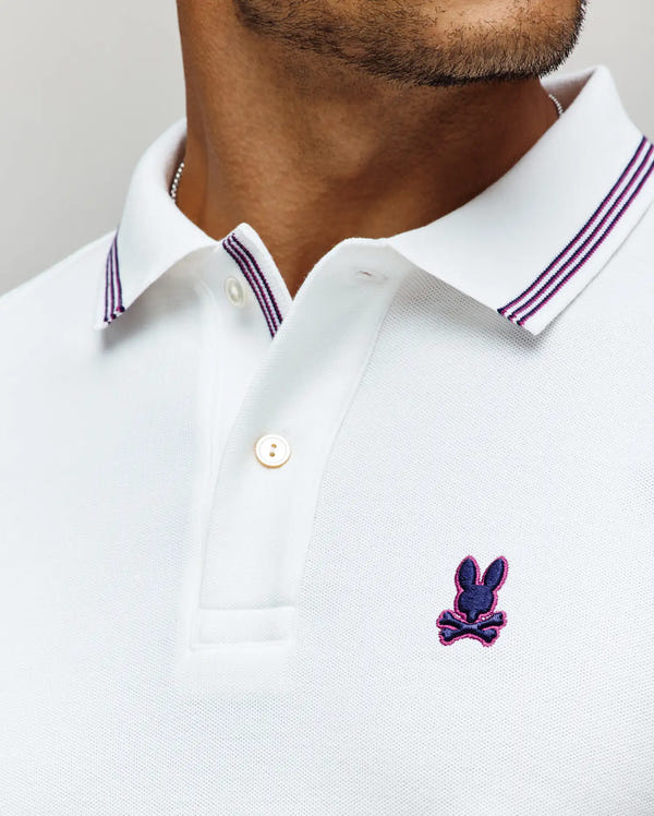 Close-up of a man wearing the Psycho Bunny MENS HART PIQUE POLO SHIRT, MT0100032, in white Pima cotton with a purple and navy striped collar and a dark blue bunny logo with pink outline on the chest.