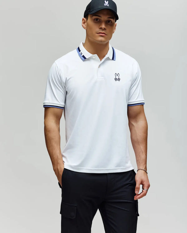 A man wearing the Psycho Bunny MENS CHRIS PERFORMANCE PIQUE POLO (MT0100038) with blue trim, black cargo pants, and a black cap stands against a plain light gray background.