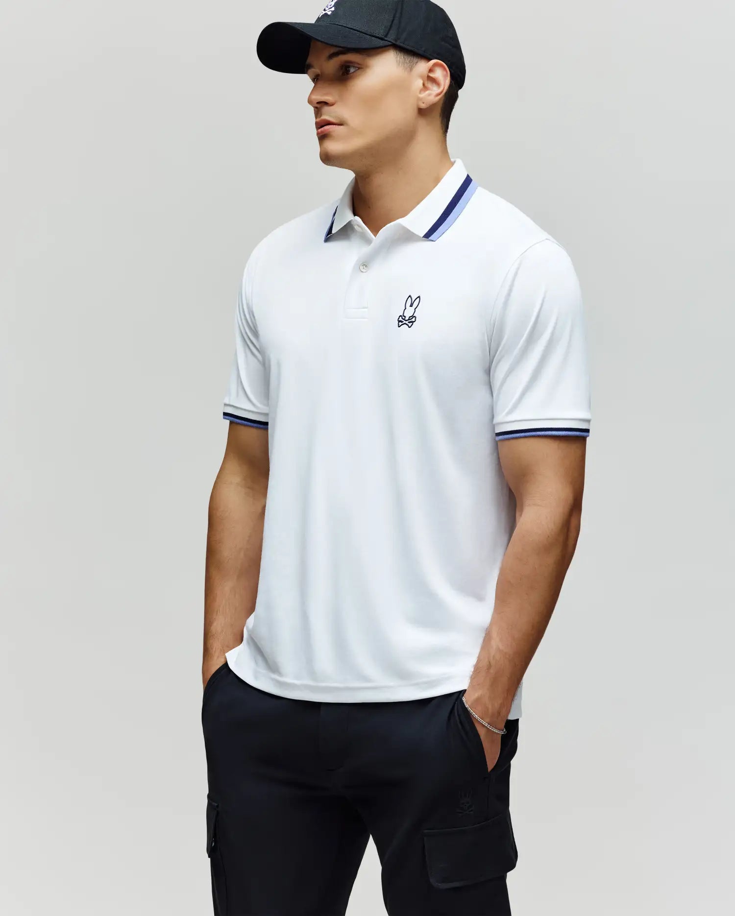 A man wearing the Psycho Bunny MENS CHRIS PERFORMANCE PIQUE POLO - MT0100038, featuring a white fabric with blue-striped collar and cuffs, pairs it with black cargo pants and a black baseball cap, standing with hands in pockets and looking to the side.