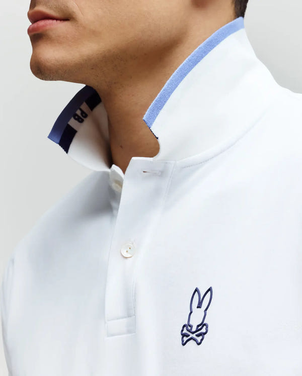 A man wears the Psycho Bunny MENS CHRIS PERFORMANCE PIQUE POLO (MT0100038) in slim fit white with blue trim and an embroidered bunny-over-bones logo on the chest. The turned-up collar reveals blue detailing inside.