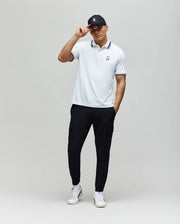 A man stands against a plain light background, wearing the Psycho Bunny MENS CHRIS PERFORMANCE PIQUE POLO (MT0100038), black cargo pants, white sneakers, and a black cap. He holds the brim of his cap with one hand in a relaxed pose.