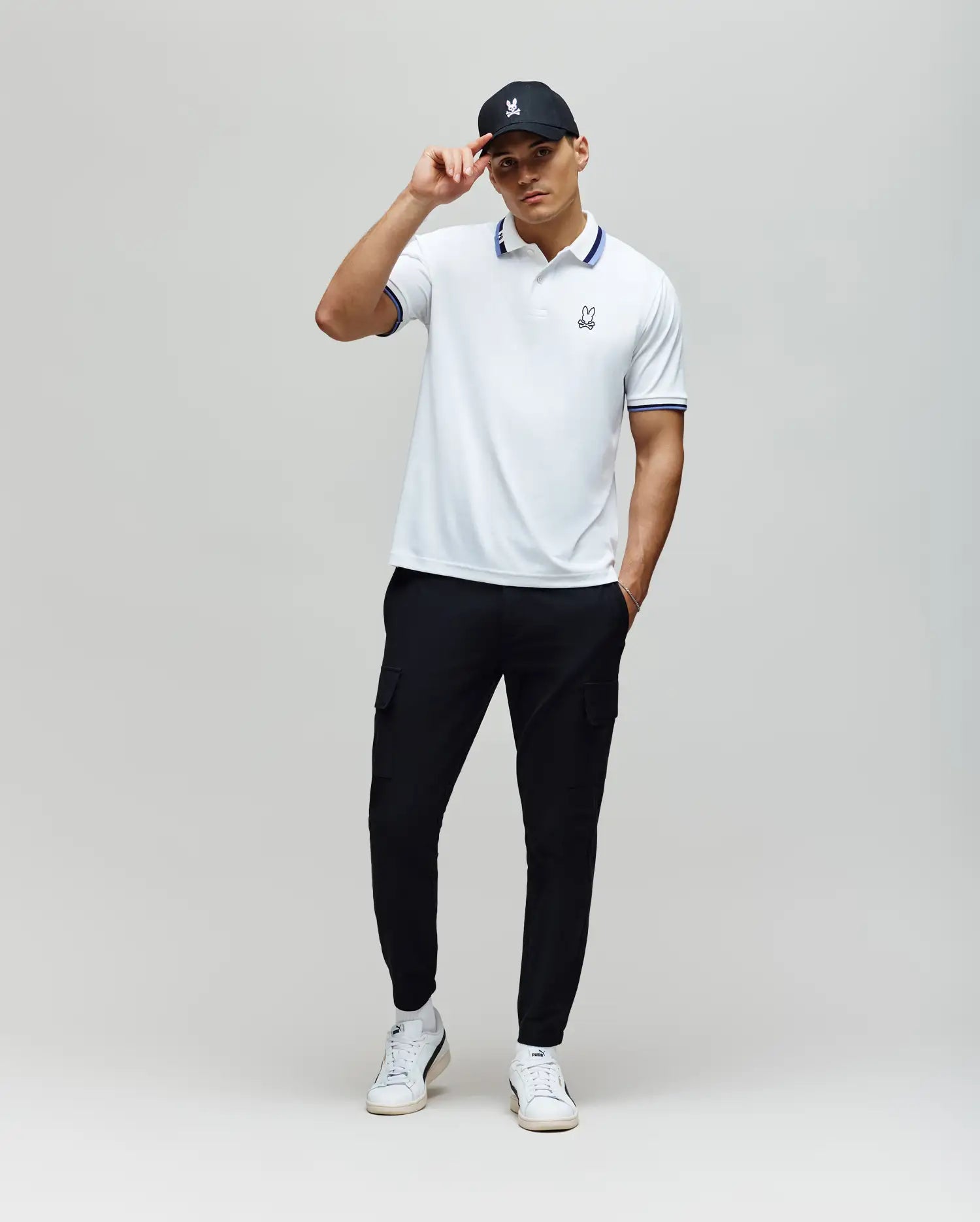 A man stands against a plain light background, wearing the Psycho Bunny MENS CHRIS PERFORMANCE PIQUE POLO (MT0100038), black cargo pants, white sneakers, and a black cap. He holds the brim of his cap with one hand in a relaxed pose.