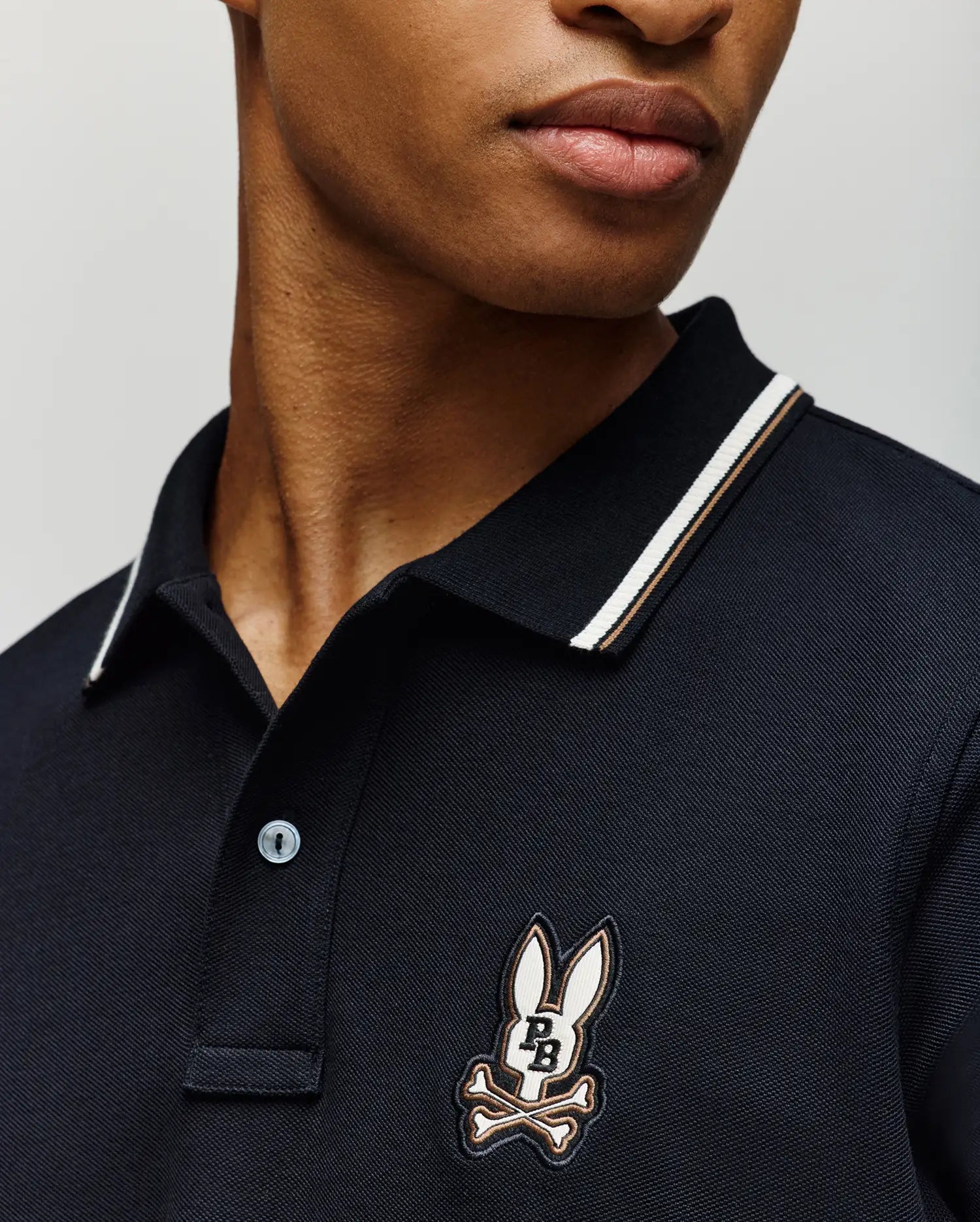 A person wears the Psycho Bunny MENS WADE PIQUE POLO (MT0100044), a dark men's shirt with a striped collar and the brand's signature cartoon bunny head above crossed bones patch on the chest. The face is partially visible against a plain background.