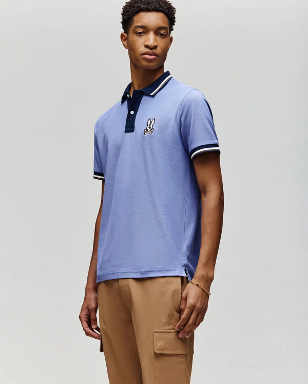 A young man stands against a light grey background wearing the MENS WADE PIQUE POLO (MT0100044) by Psycho Bunny in light blue, featuring an embroidered bunny logo on the chest, paired with tan cargo pants.