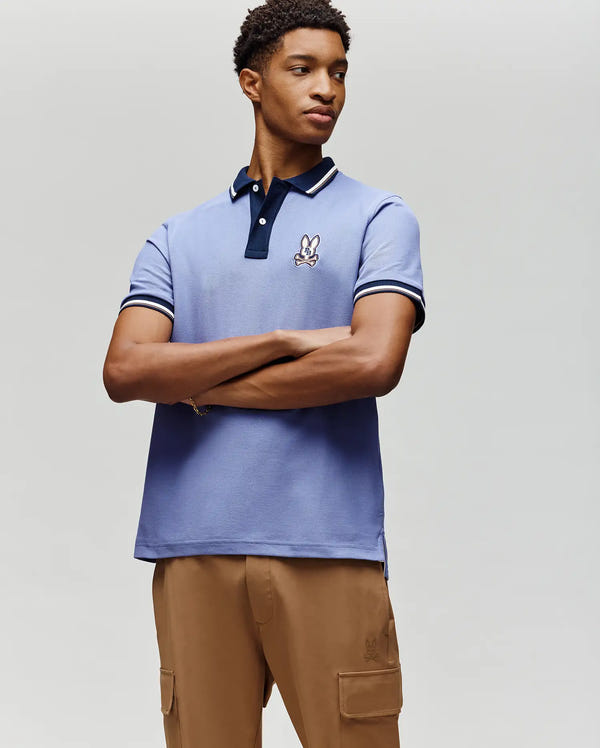A young man stands with arms crossed in front of a plain light background, wearing the Psycho Bunny MENS WADE PIQUE POLO (MT0100044) in light blue with an embroidered bunny logo on the chest and tan cargo pants.