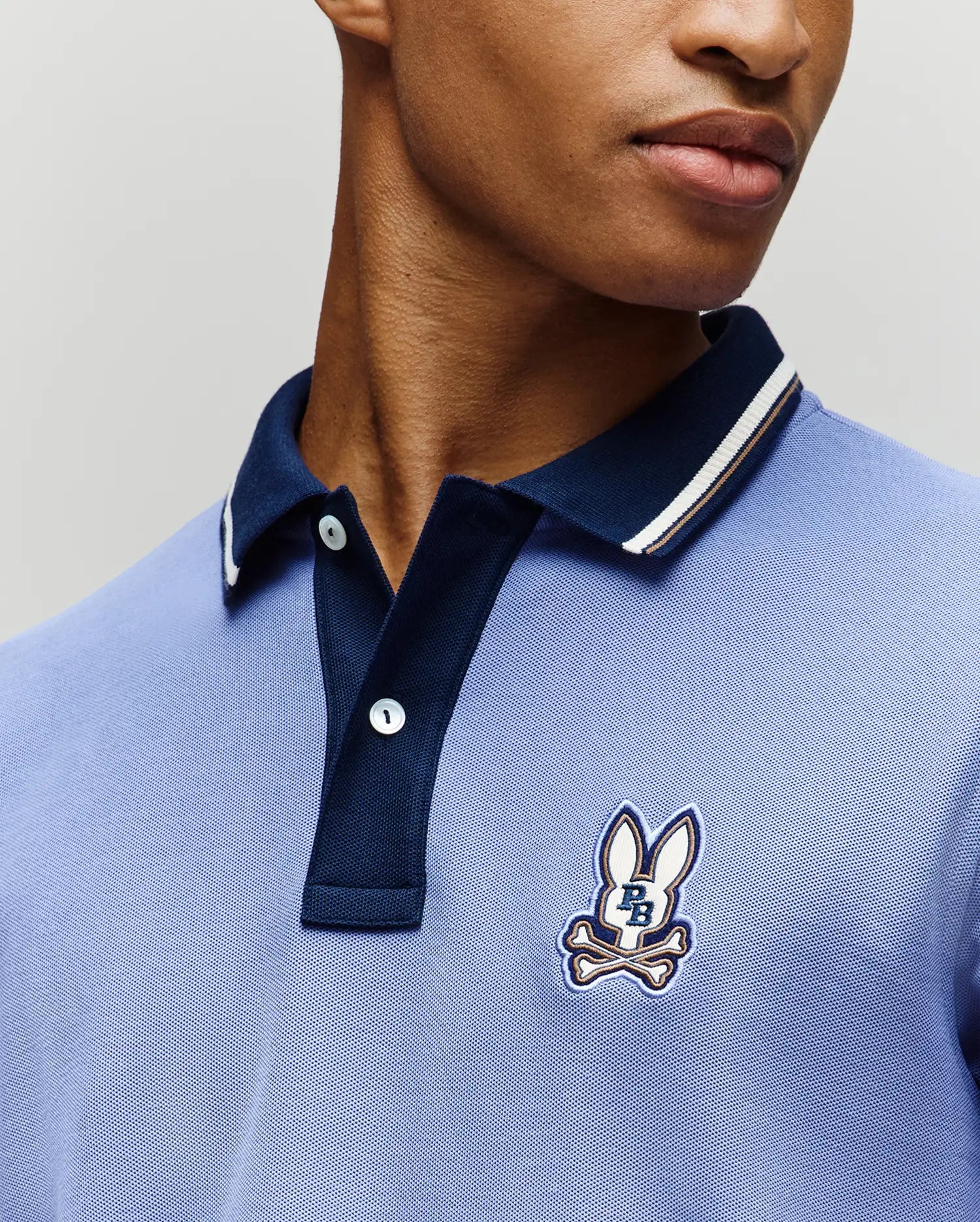 A person wears the Psycho Bunny MENS WADE PIQUE POLO (MT0100044), a light blue Pima cotton polo with a dark blue collar, colorblock tipping, and an embroidered bunny and crossbones logo. Only the lower face and upper torso are visible.