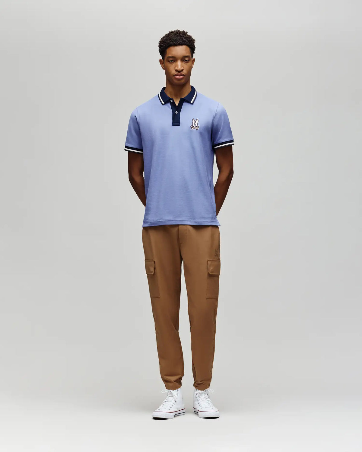 A man stands against a plain gray background, wearing the Psycho Bunny MENS WADE PIQUE POLO - MT0100044 in light blue, paired with tan cargo jogger pants and white sneakers. His hands are behind his back as he looks straight ahead.