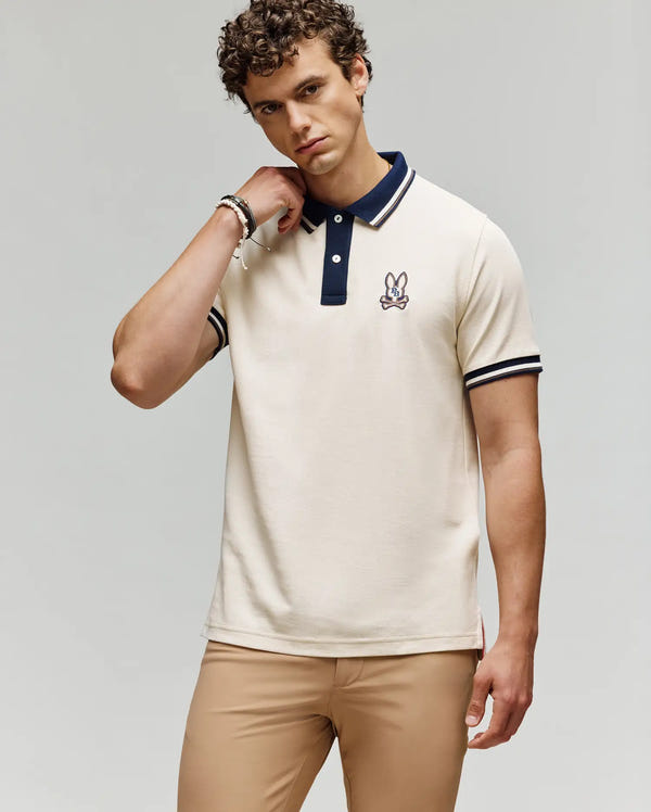A man with curly hair wears the Psycho Bunny MENS WADE PIQUE POLO - MT0100044, featuring a black collar and embroidered bunny logo, paired with beige pants against a plain light background.
