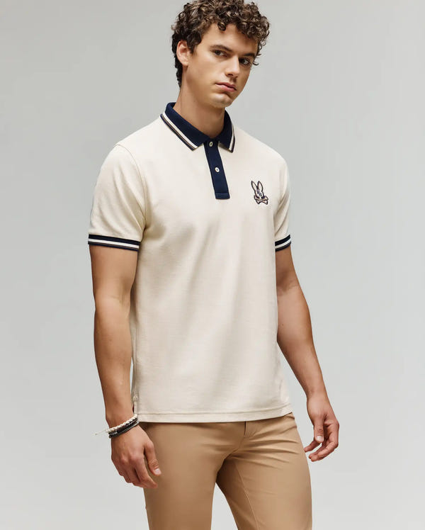 A man with curly hair wears the Psycho Bunny MENS WADE PIQUE POLO - MT0100044, a cream Pima cotton polo with black trim and an embroidered bunny logo, paired with tan pants against a plain light gray background.