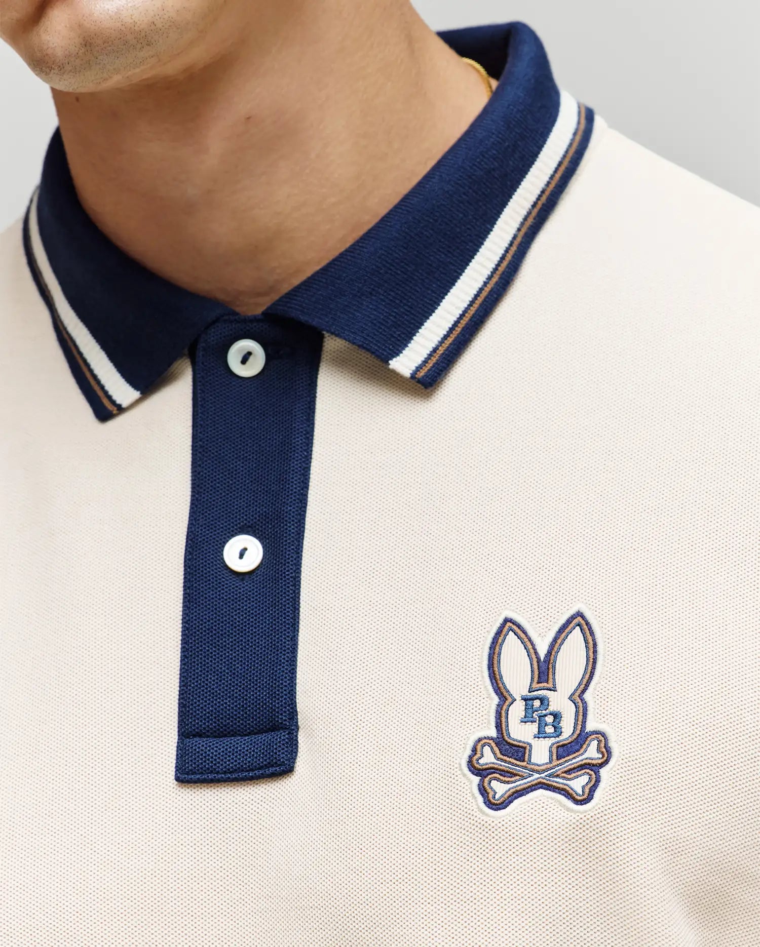 A close-up shows a person wearing the Psycho Bunny MENS WADE PIQUE POLO (MT0100044), a colorblock style with a navy collar, buttons, and an embroidered bunny logo above crossed bones plus "P B" on the left chest.